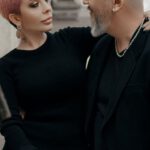 Communication - Elegant short haired woman in black dress embracing stylish bald middle aged man while standing together near old stone building in city