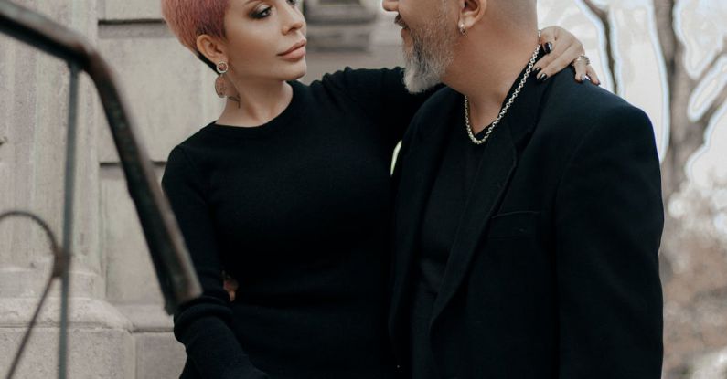 Communication - Elegant short haired woman in black dress embracing stylish bald middle aged man while standing together near old stone building in city
